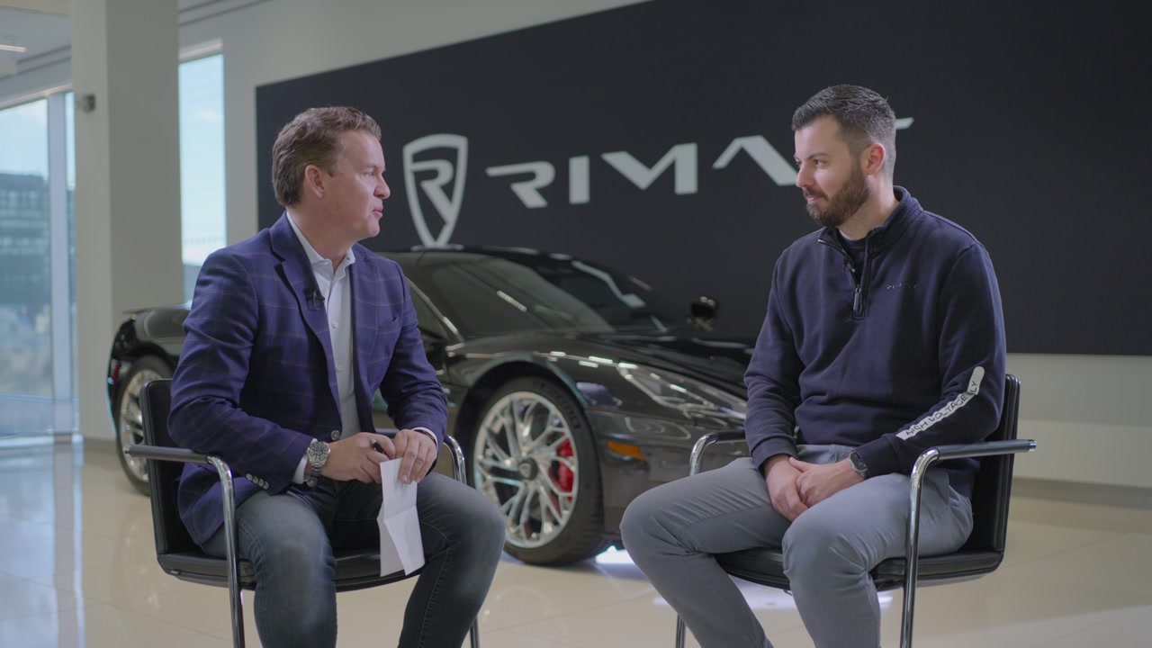 Two men talking in chairs in front of a sports car and a Rimac sign