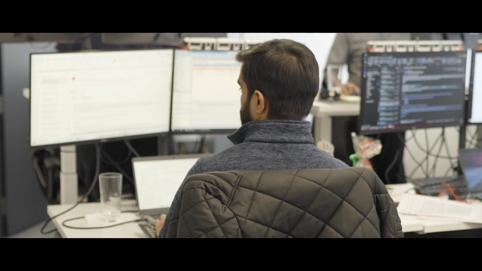 A man at a desk with large monitors and a laptop in an open office, a colleague standing behind