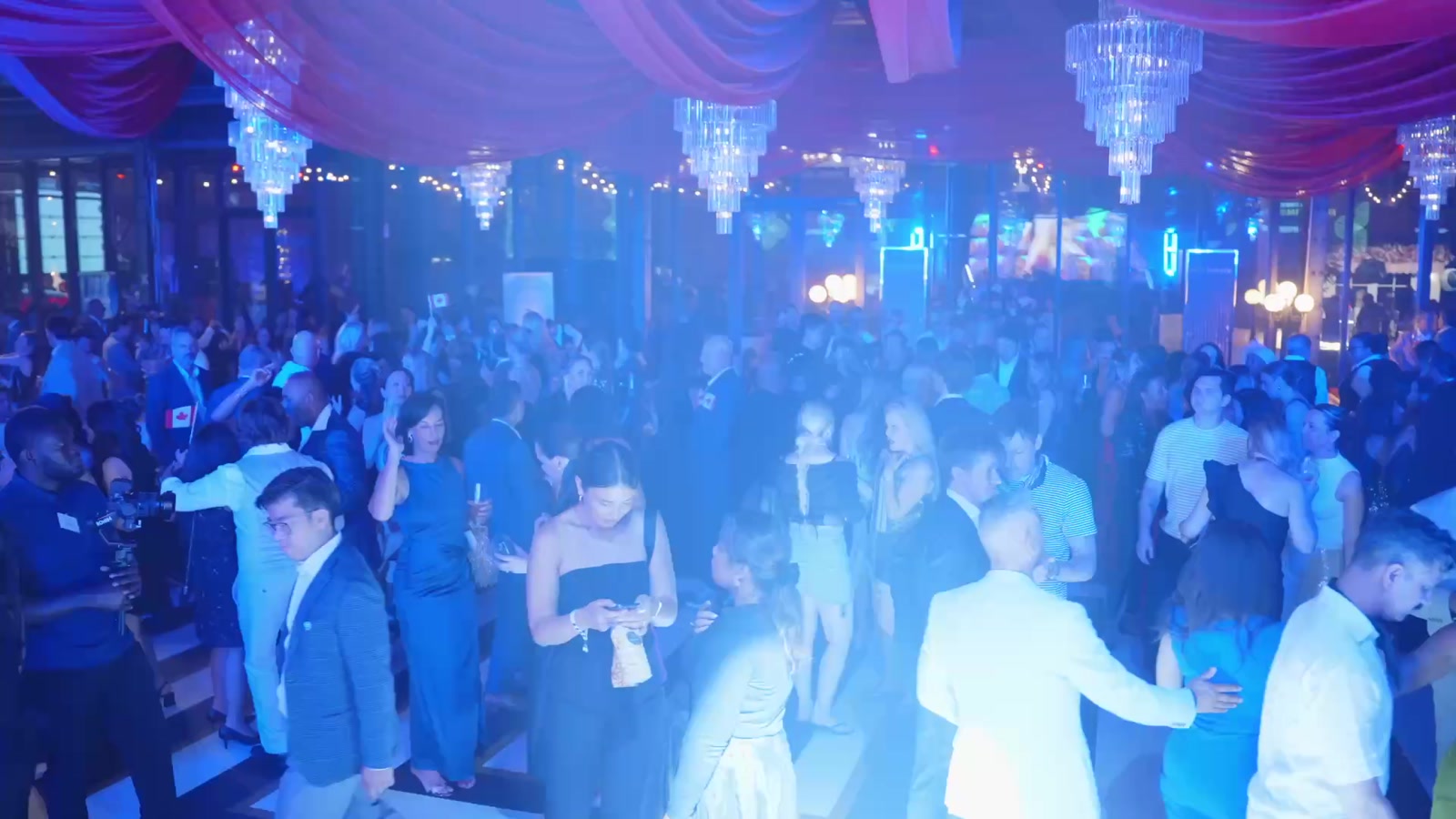 A crowded dance floor under chandeliers and blue light at a gala