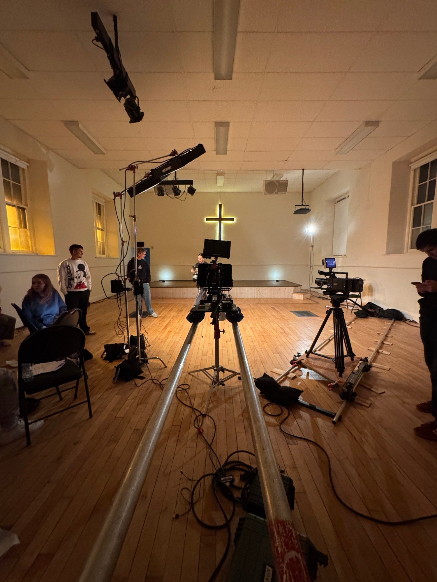 Lights, cameras and a dolly track set up in a hall with a lit cross on the wall