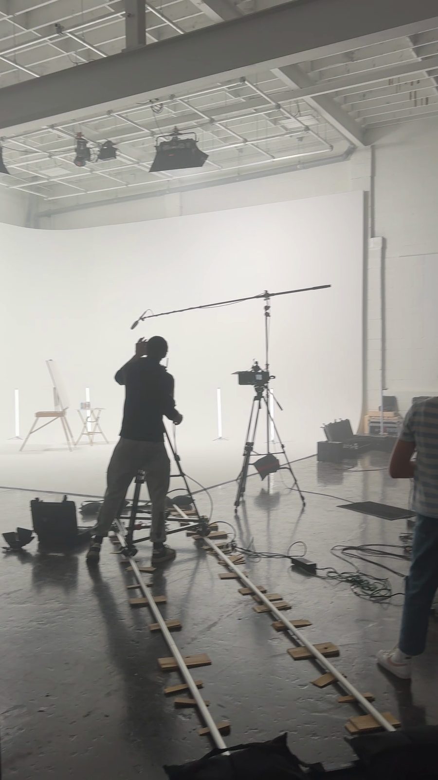 A camera operator on a dolly track in a large white studio, with a boom microphone, a second camera and lights overhead