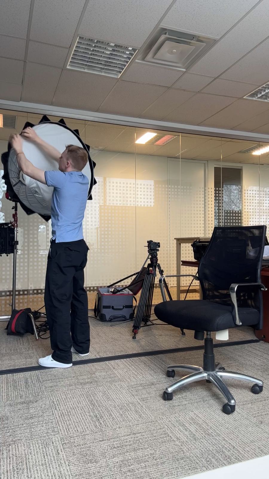 A crew member fitting a large round softbox to a light stand in an office with frosted glass walls, a camera on a tripod beside an empty desk chair