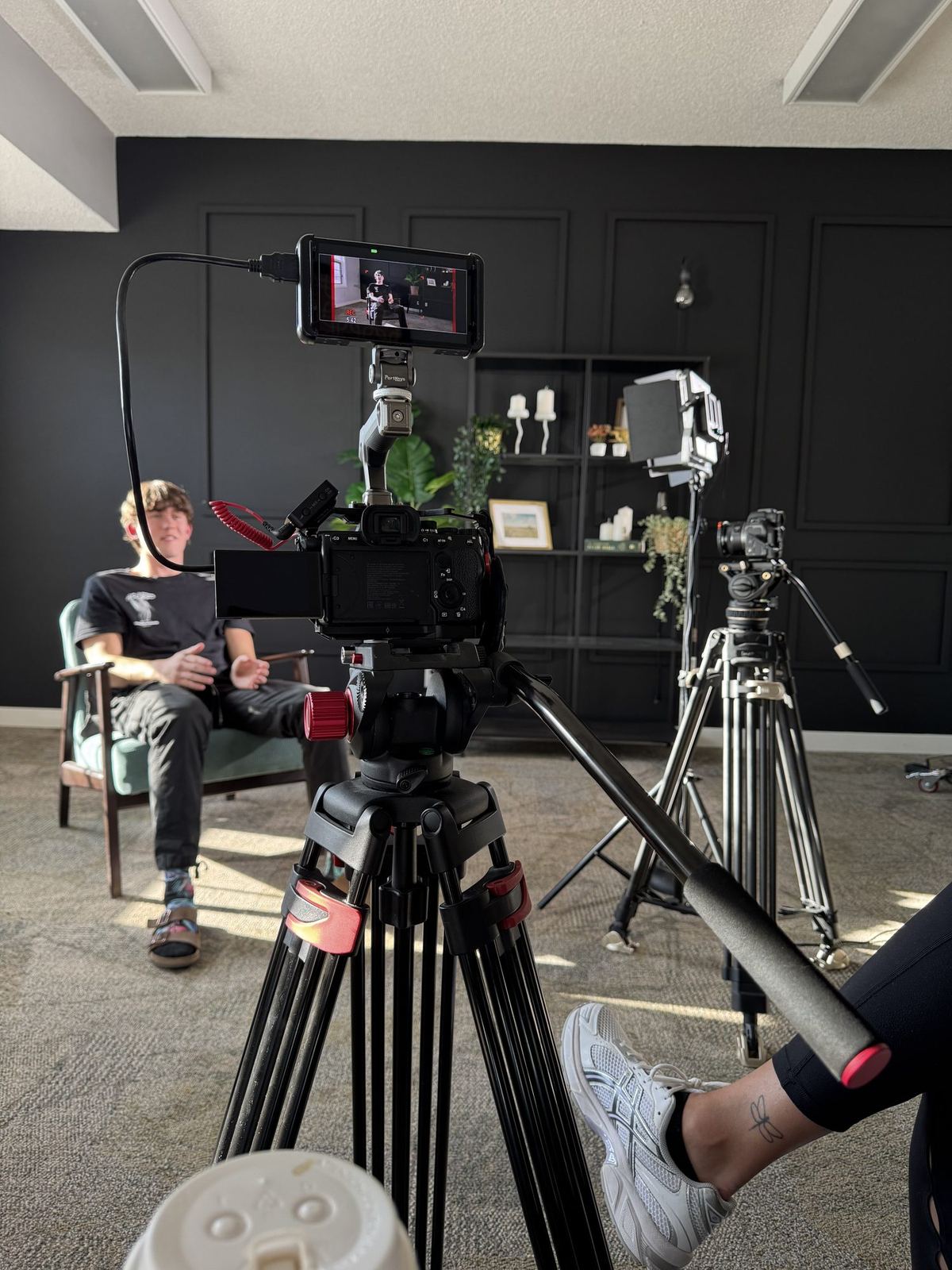 An interview set: a camera and monitor on a tripod facing a seated speaker in a dark-panelled room, with a second camera and a light behind