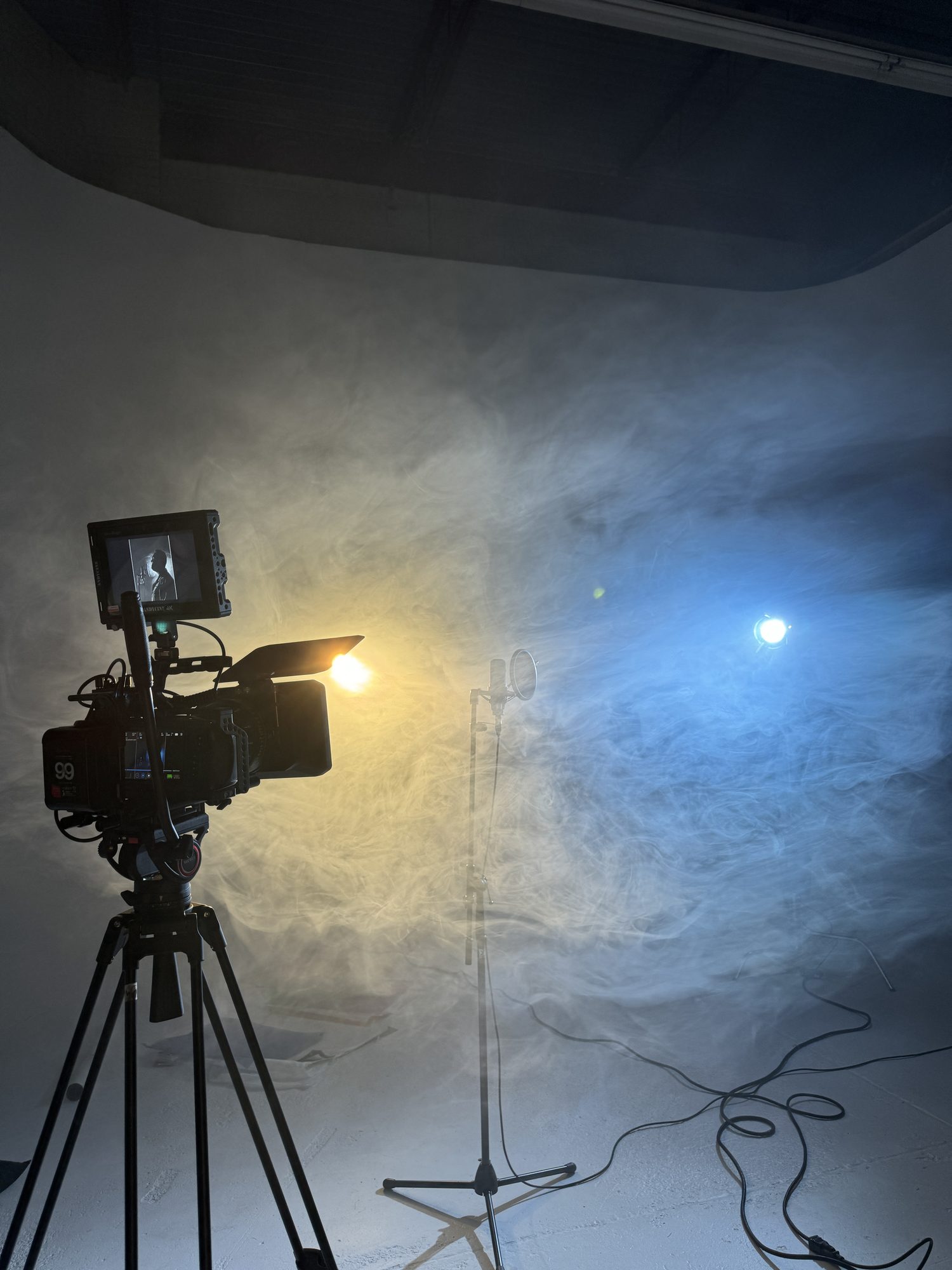 A cinema camera on a tripod and a microphone stand in a hazy studio lit by one warm and one blue light